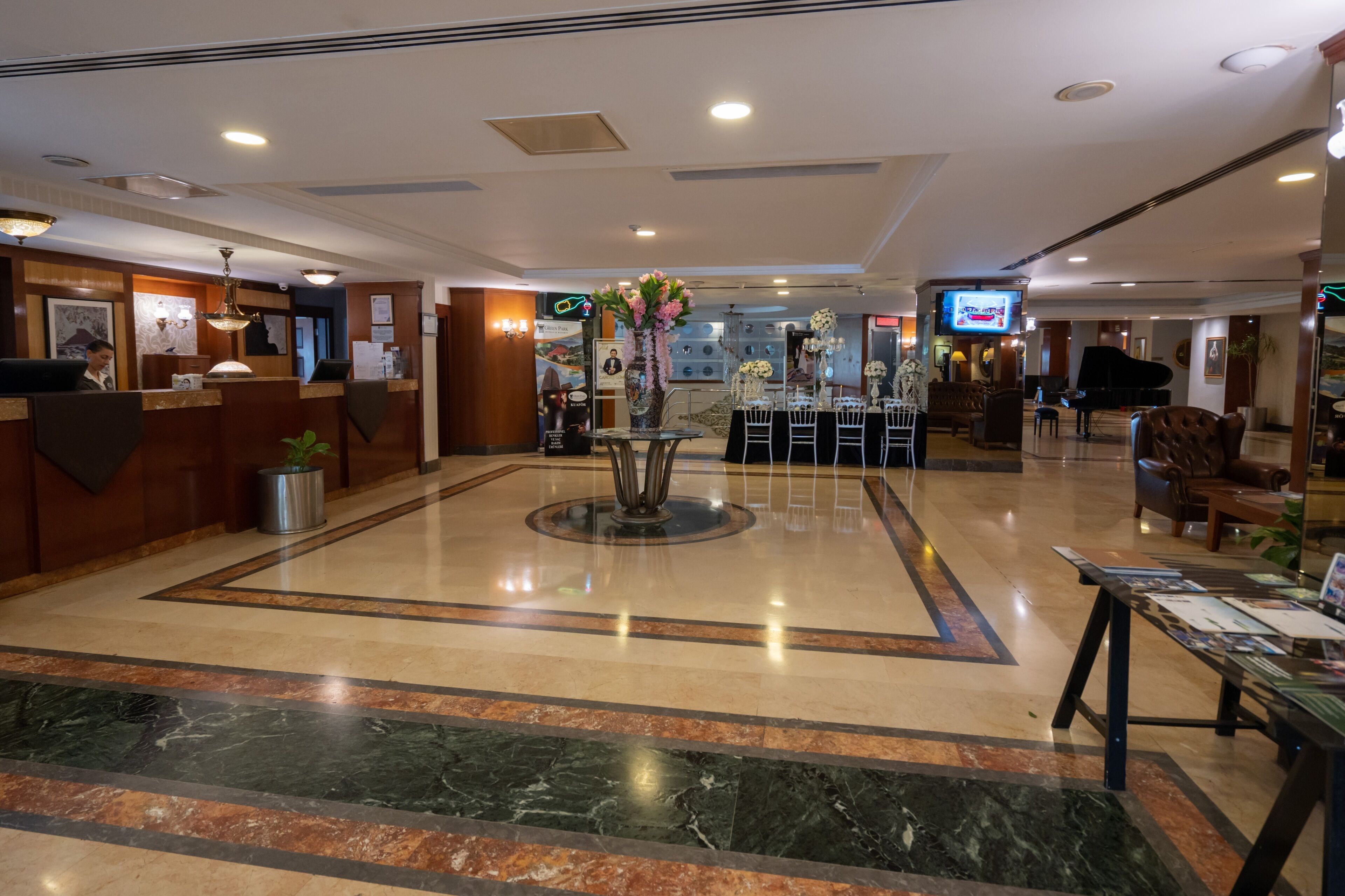 A hotel lobby with a reception desk, a central decorative area with flowers, a piano, and seating arrangements.