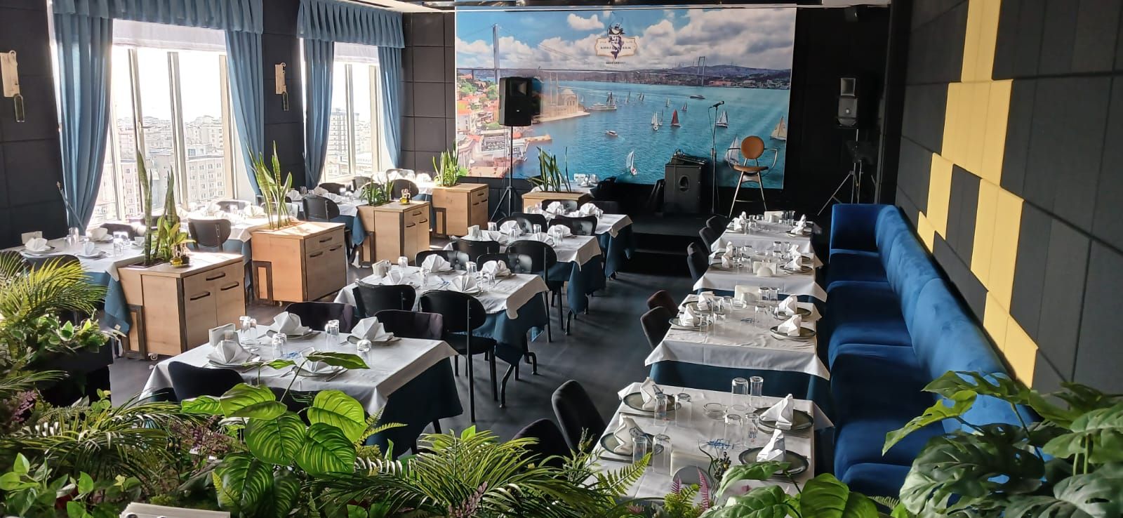 En restaurang med dukade bord, en väggmålning som föreställer ett kustlandskap och krukväxter.