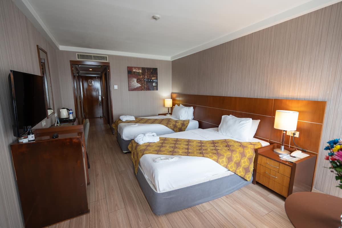 Quarto de hotel com duas camas, uma escrivaninha, uma televisão e um banheiro visível por uma porta aberta.