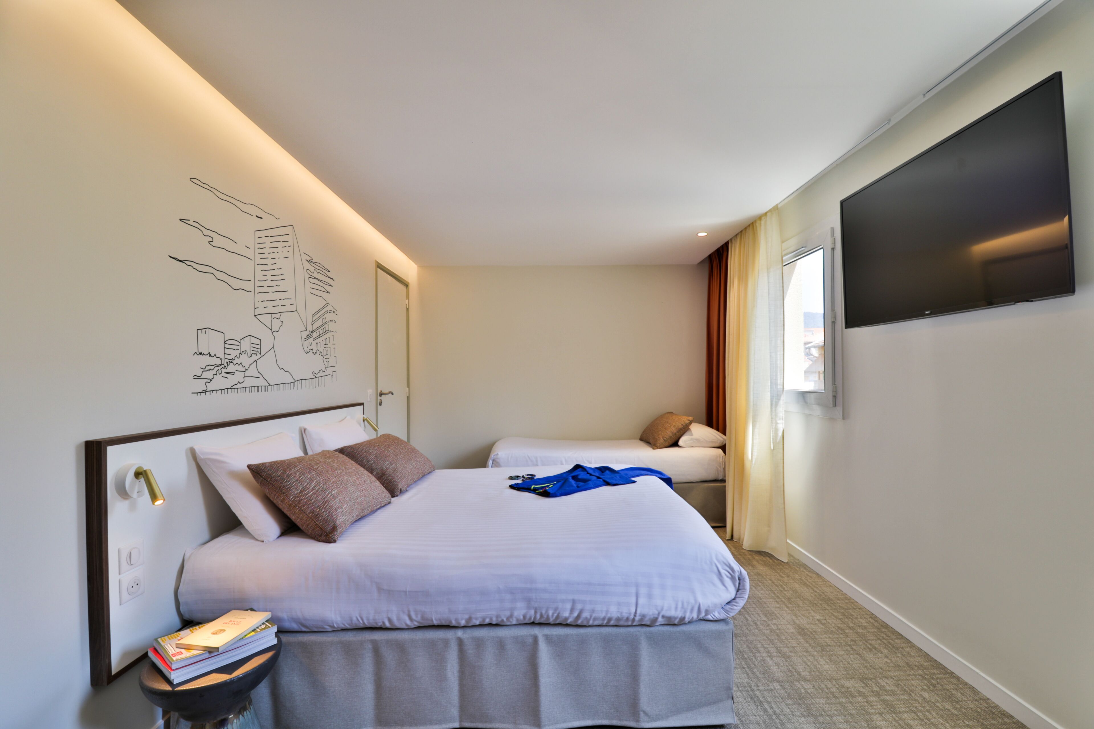 A hotel room with two beds, a flat-screen TV, a bedside table with books, and a wall-mounted artwork.