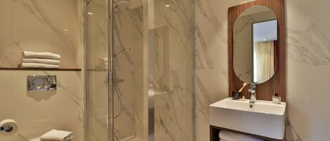 A modern bathroom with a glass-enclosed shower, a marble-tiled wall, a white sink with a wooden vanity, and a toilet with stacked towels.