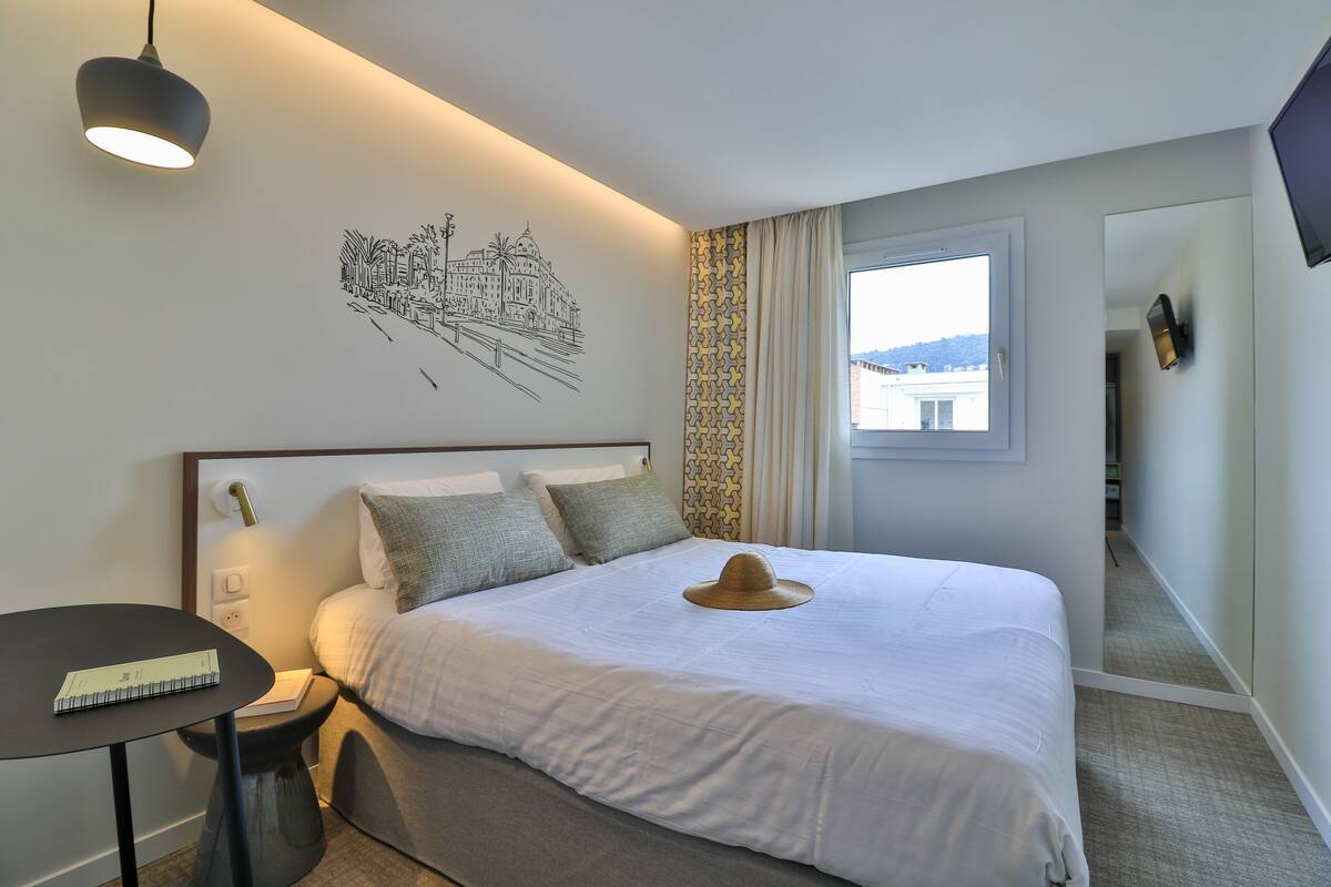A neatly made bed with a hat on it, a bedside table with a book, a wall art of a building, a window with curtains, and a TV on the wall.