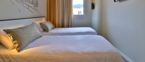 A hotel room with two beds, a window, a wall-mounted TV, and a cityscape mural.