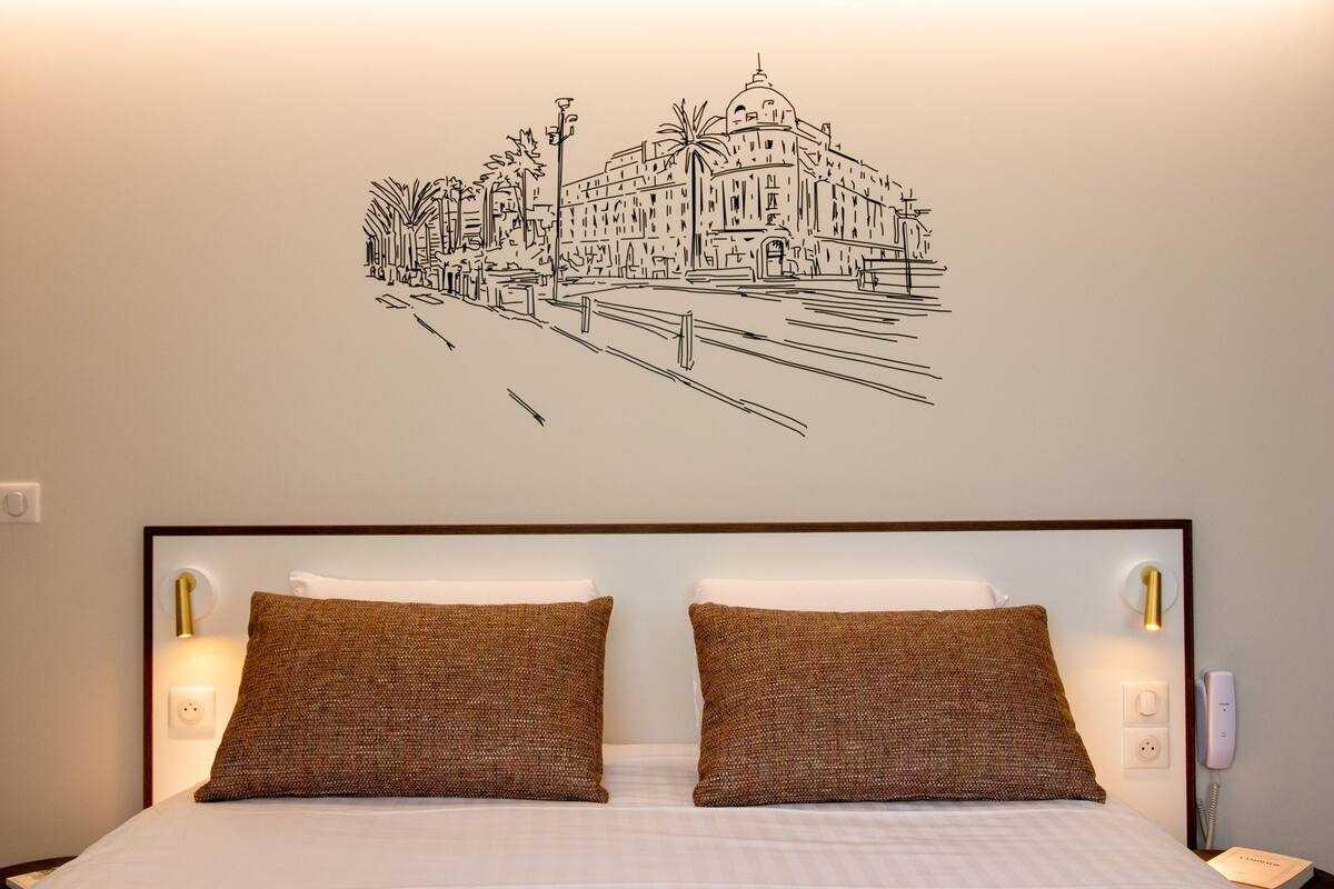 A hotel room with a bed, two pillows, a wall mural, and a bedside table with a lamp.