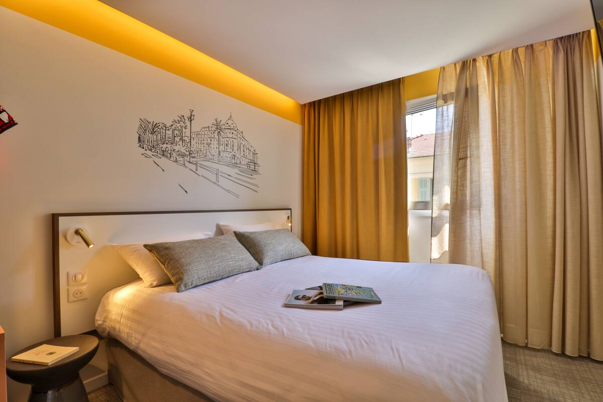 A hotel room with a large bed, a nightstand, a wall art of a cityscape, and a window with curtains.