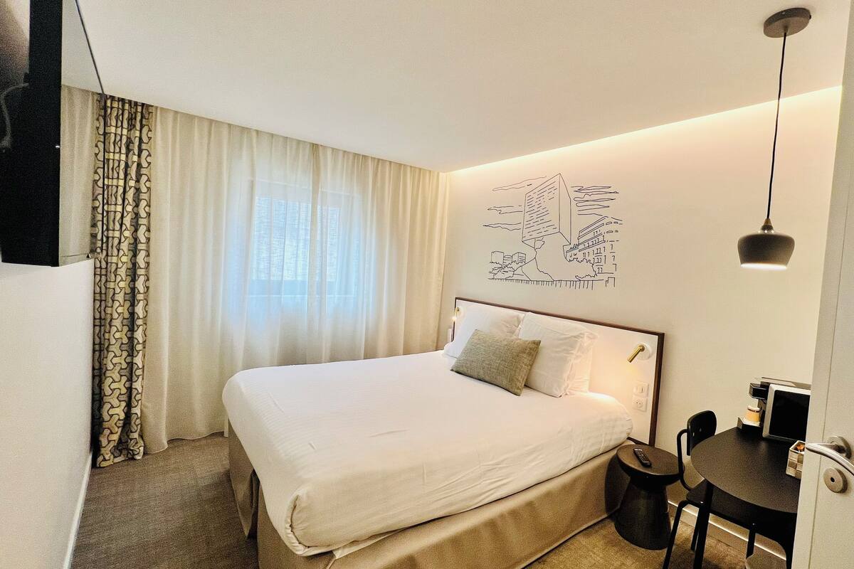 A neatly made bed with a white comforter and pillows, a small round table with a remote control, and a wall-mounted light fixture.