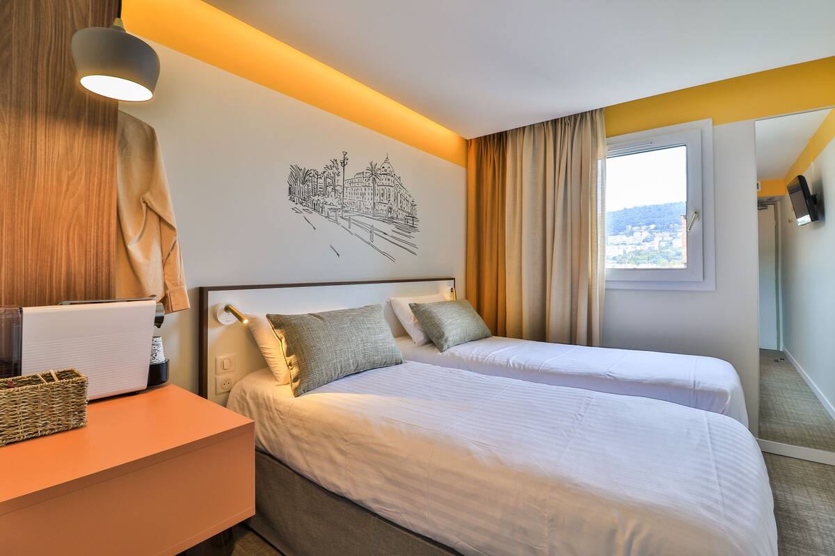 A hotel room with two beds, a desk with a lamp, a wall art of a cityscape, and a window with curtains.