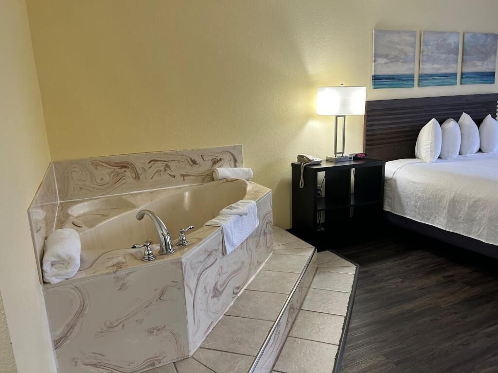 Habitación de hotel con una bañera de mármol, cama con ropa blanca, mesita de noche con lámpara y un cuadro en la pared.