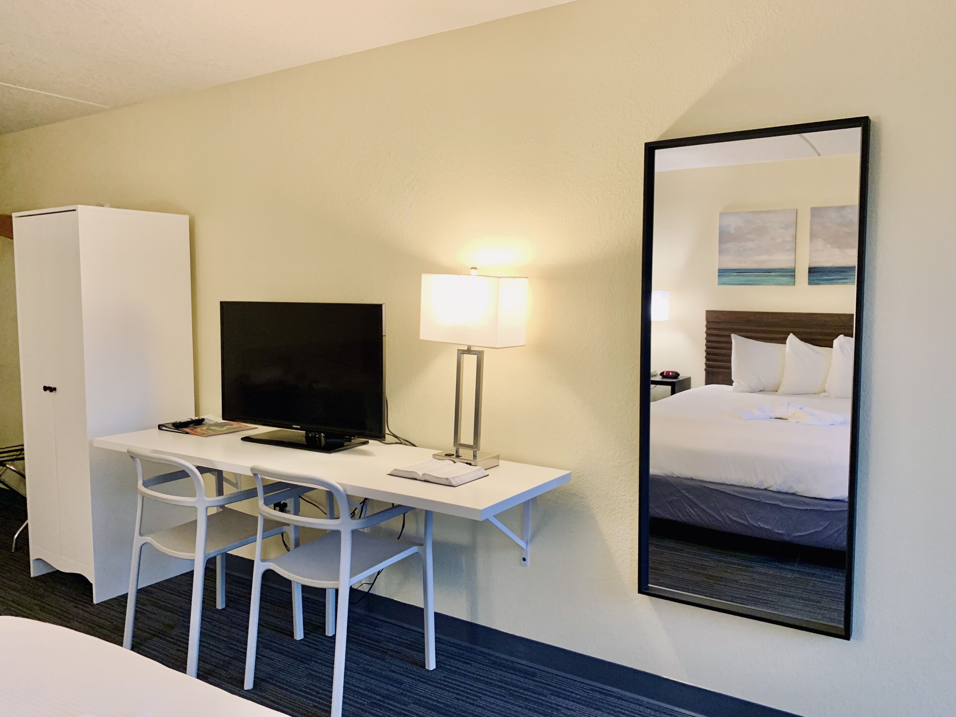 A hotel room with a desk, chair, television, lamp, and a large mirror reflecting a bed.