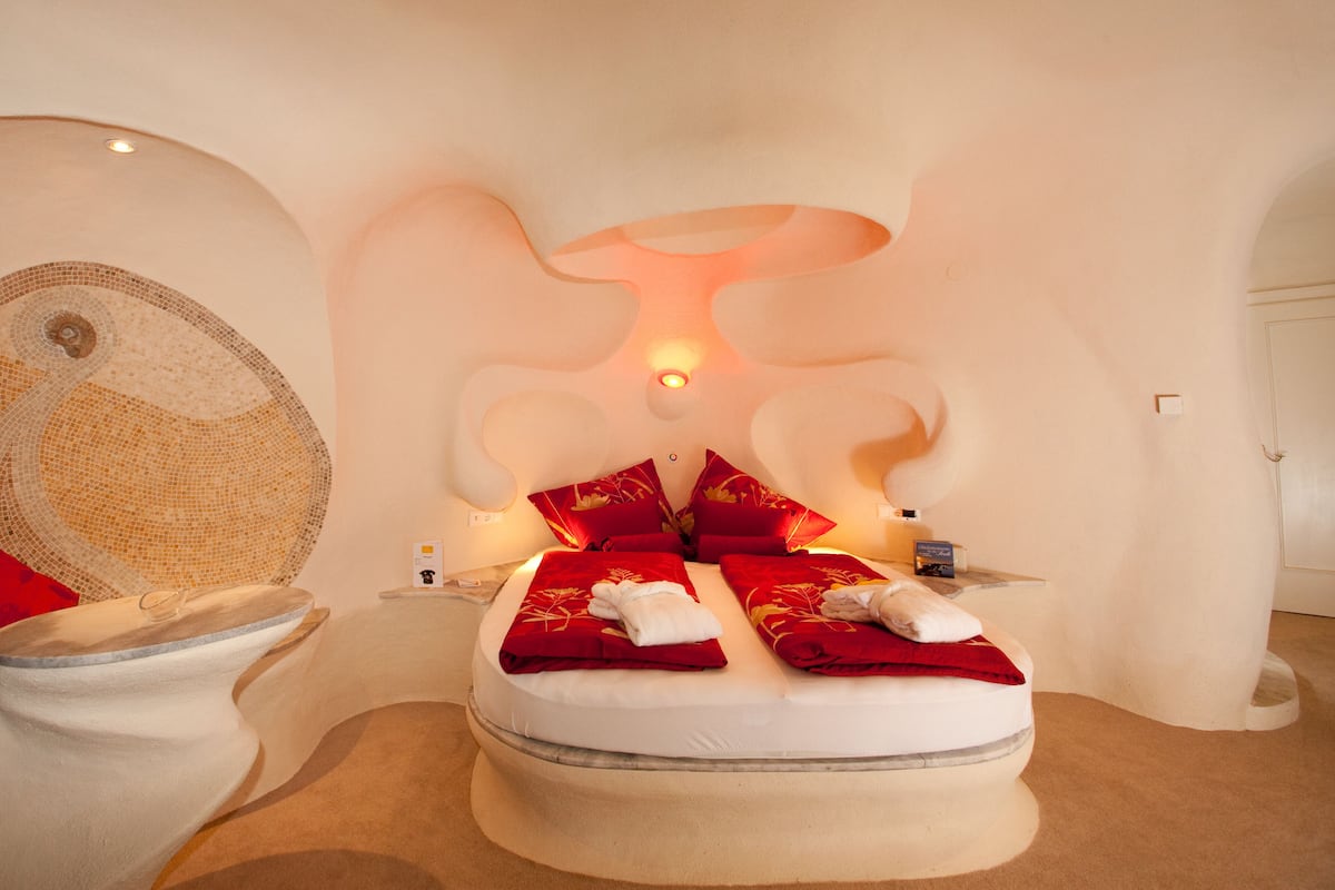 A uniquely designed bedroom with a circular mosaic wall, a bed with red bedding, and a modern light fixture above the bed.
