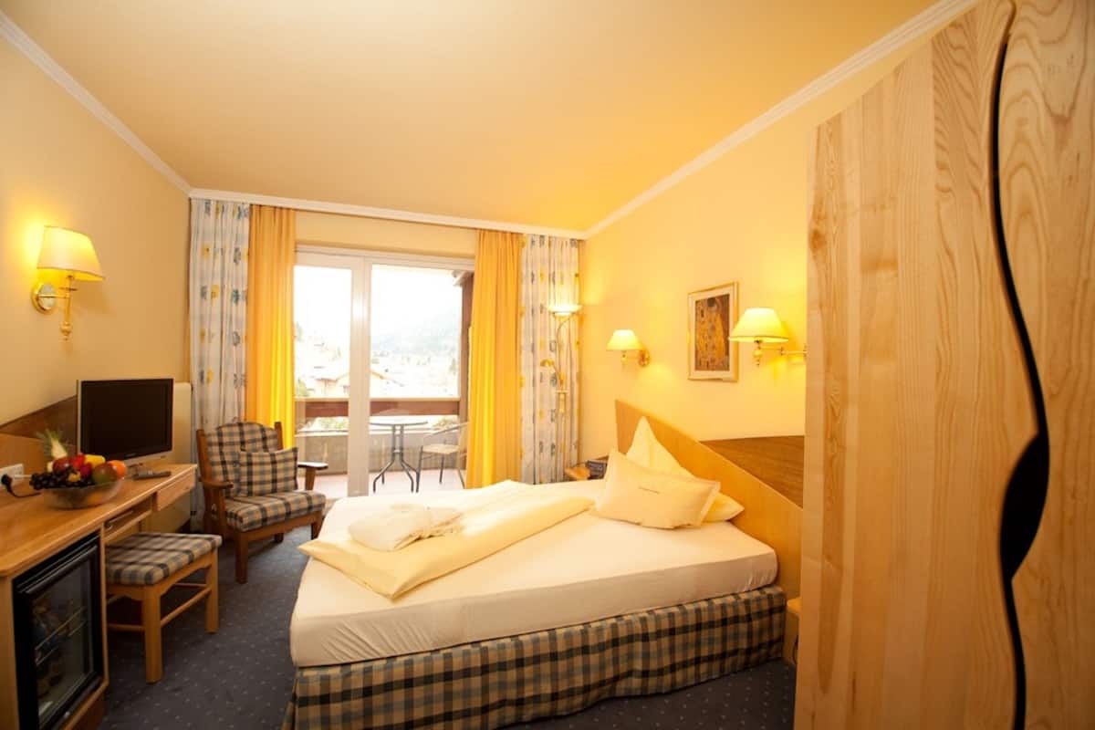 A hotel room with a bed, a desk with a computer, a chair, a dining table, and a balcony with curtains.