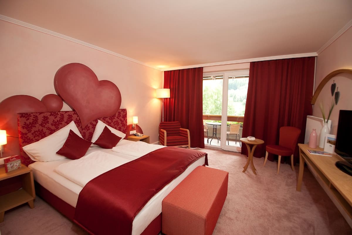A hotel room with a large bed, a desk with a chair, a small table, and a balcony with red curtains.