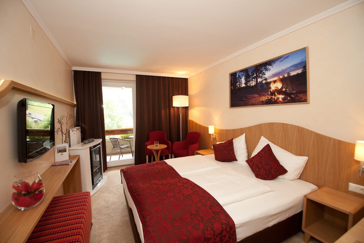 A hotel room with a large bed, a desk with a TV, a small table with chairs, a fireplace, and a painting of a campfire.