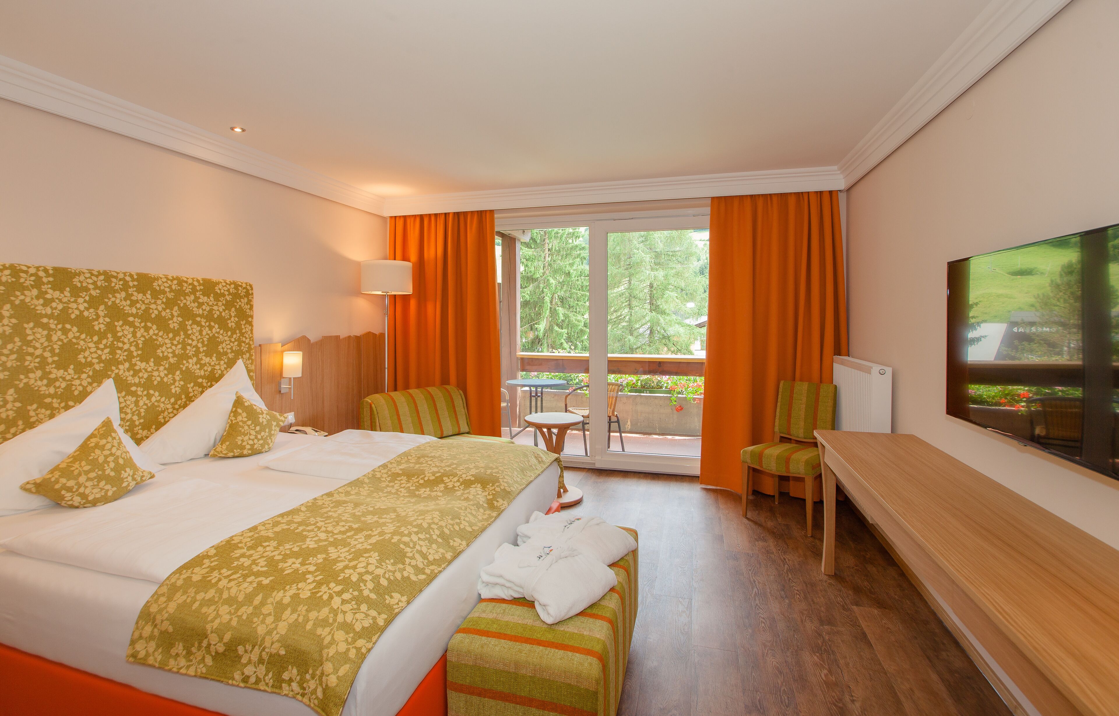 Habitación de hotel con una cama grande, un banco, un televisor, una mesa con sillas y un balcón con cortinas naranjas.