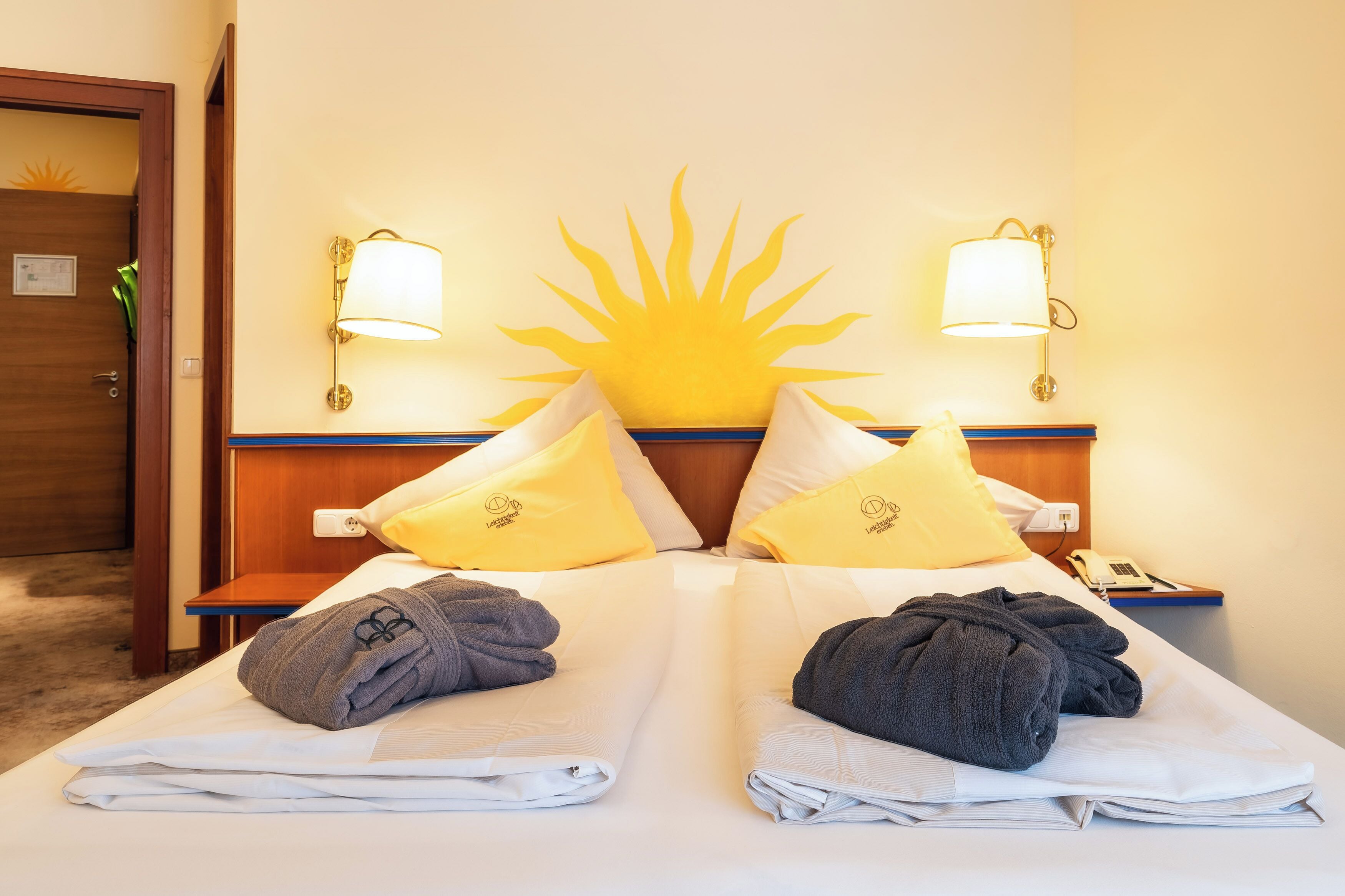 A hotel room with a bed, two pillows, a nightstand with a phone, and wall-mounted lamps.
