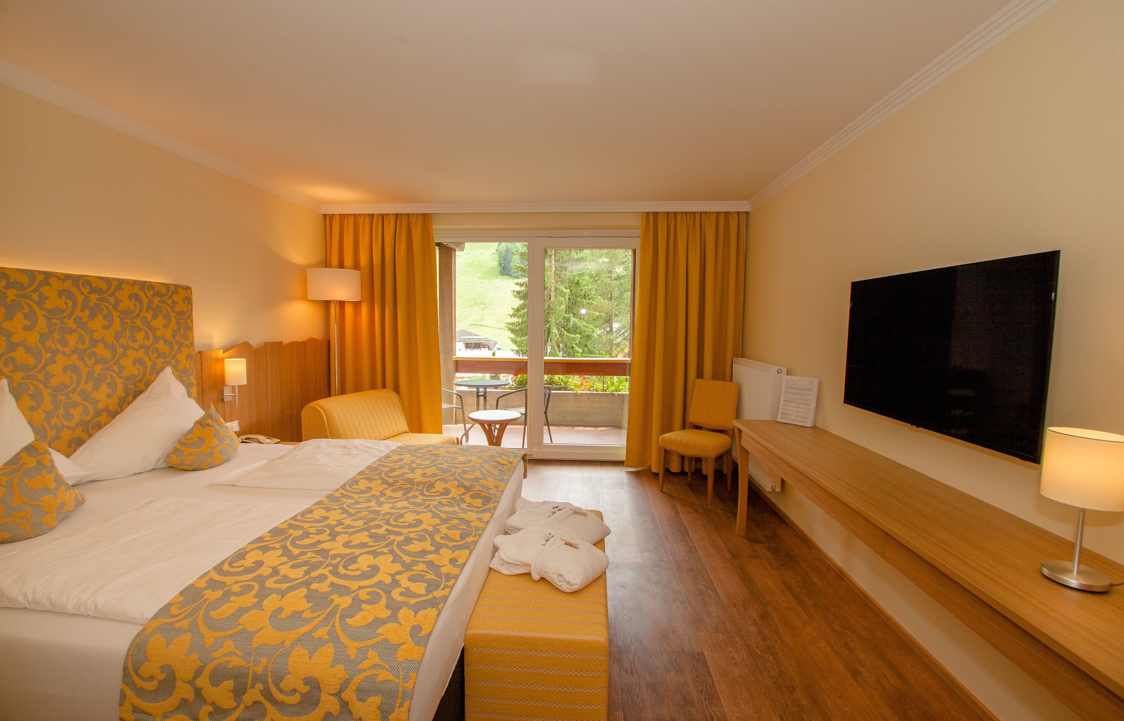 Ein Hotelzimmer mit einem großen Bett, einem Flachbildfernseher, einem kleinen Tisch und einem Balkon mit gelben Vorhängen.