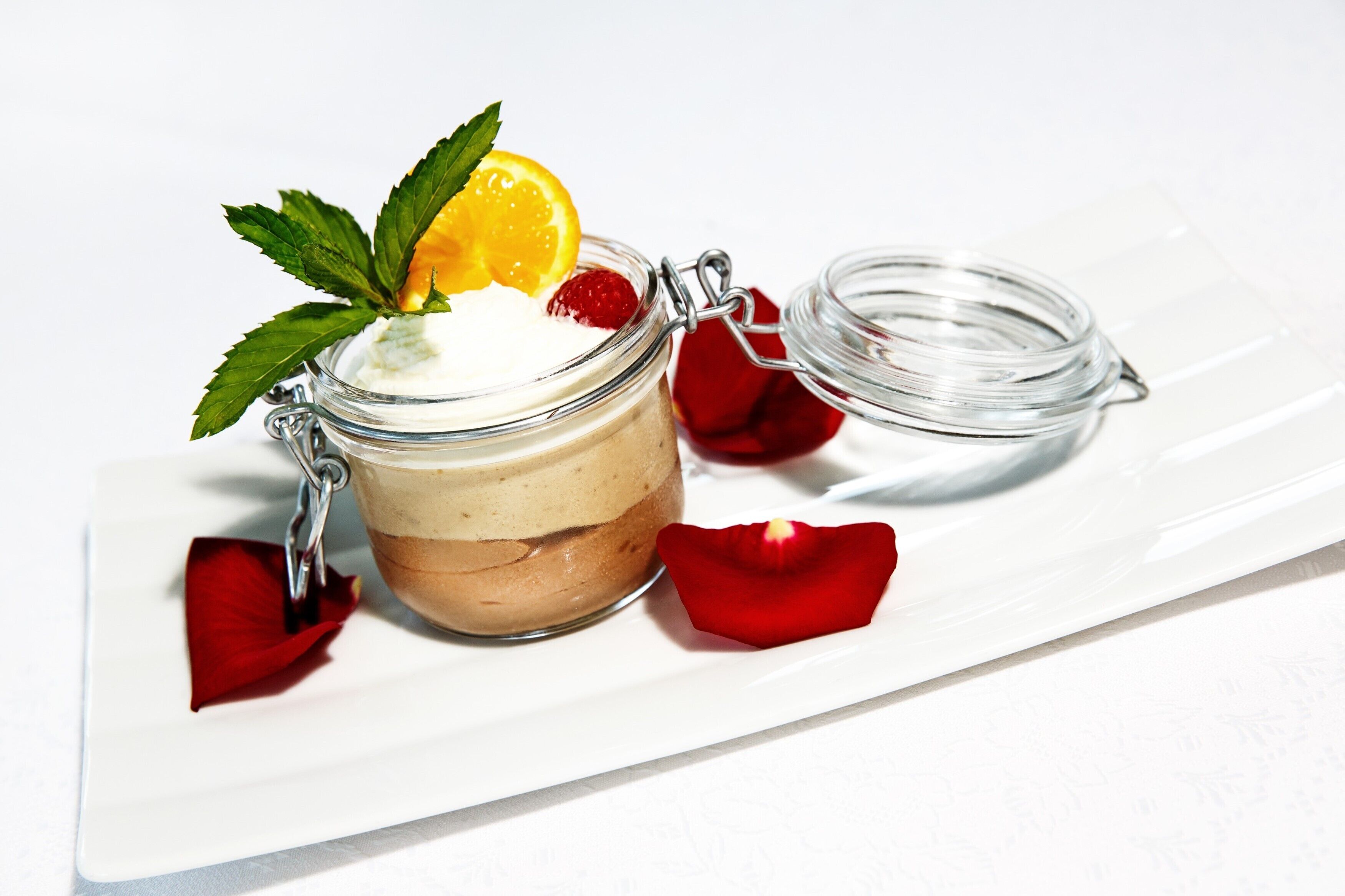 Postre en un tarro de cristal con cierre metálico, adornado con una hoja de menta, rodaja de naranja y una fresa, sobre un plato blanco con pétalos de rosa roja.