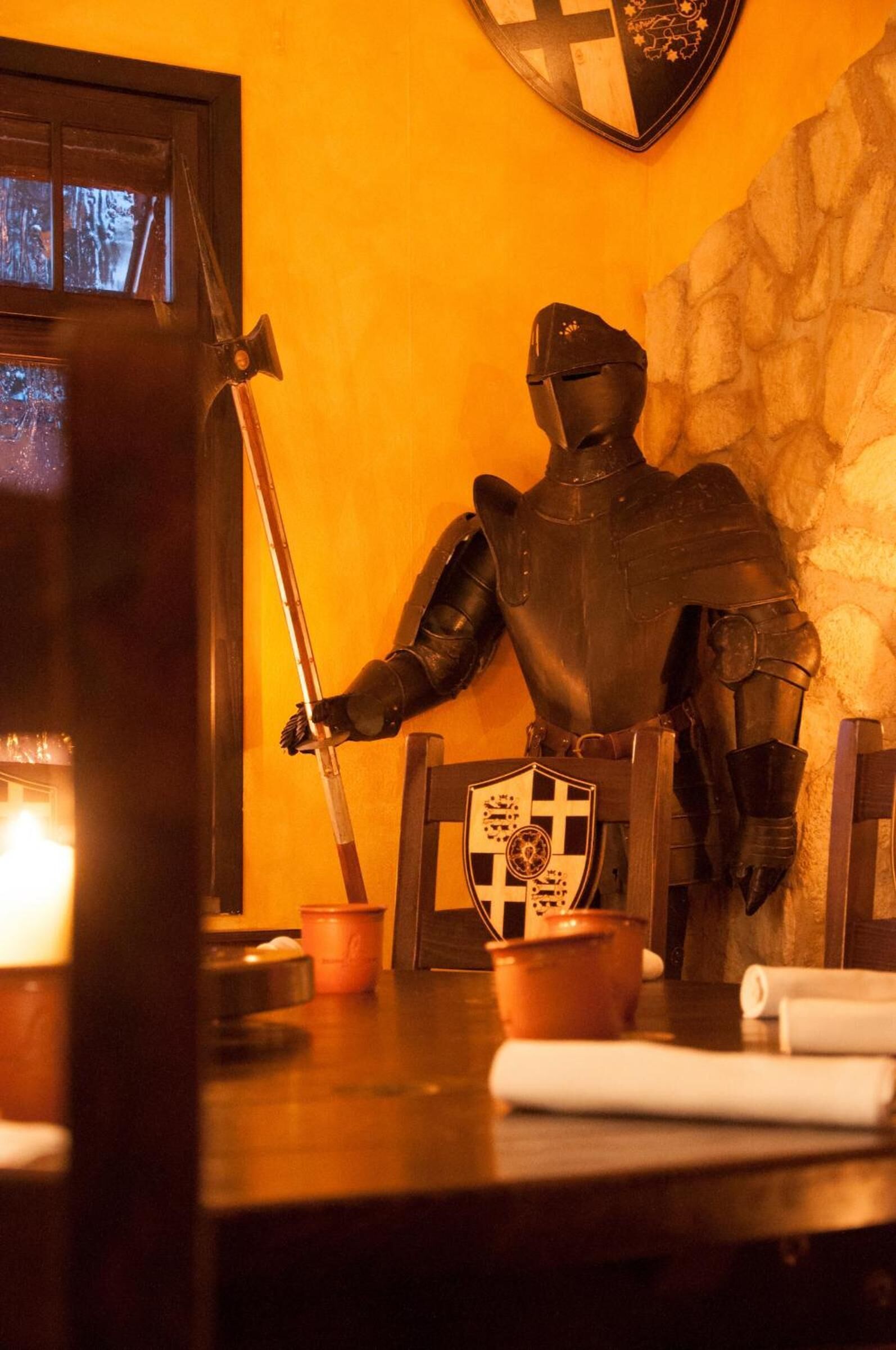 A knight's statue in armor holding a sword, a shield with a cross, and a wooden chair.