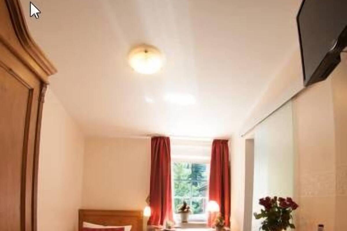 Il s’agit d’une pièce de taille modeste, comprenant un lit, un bureau et une chaise. On y trouve une fenêtre avec des rideaux, une petite table sur laquelle sont posés un vase et une bouteille, ainsi qu’un téléviseur fixé au mur.