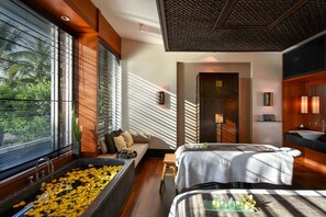 A modern hotel room with a large window, a bathtub filled with yellow flowers, a sofa, a bed, and a bathroom with a sink and mirror.