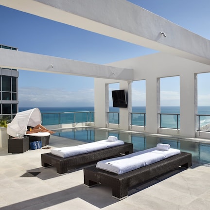 A rooftop infinity pool with two sun loungers, a flat-screen TV, and a city view.