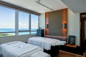 A modern hotel room with two beds, a large window with a view of the ocean, a wooden panel wall, and a small bedside table with a plant.