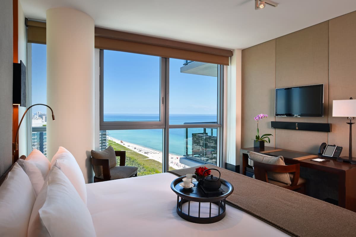 Ein Hotelzimmer mit großem Fenster und Blick auf Strand und Meer, ein Bett mit weißer Bettwäsche, ein Schreibtisch mit Computer sowie ein flachbildschirmfernseher an der Wand.