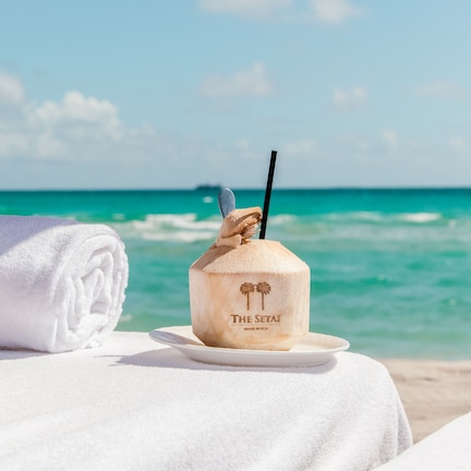 A beachside setting with a coconut drink on a plate, a rolled towel, and an umbrella.