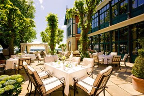 An outdoor dining area with tables and chairs, surrounded by trees and a building with large windows.