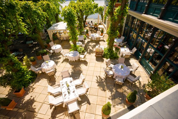 Un patio al aire libre con mesas redondas y sillas blancas, rodeado de plantas en macetas y árboles.