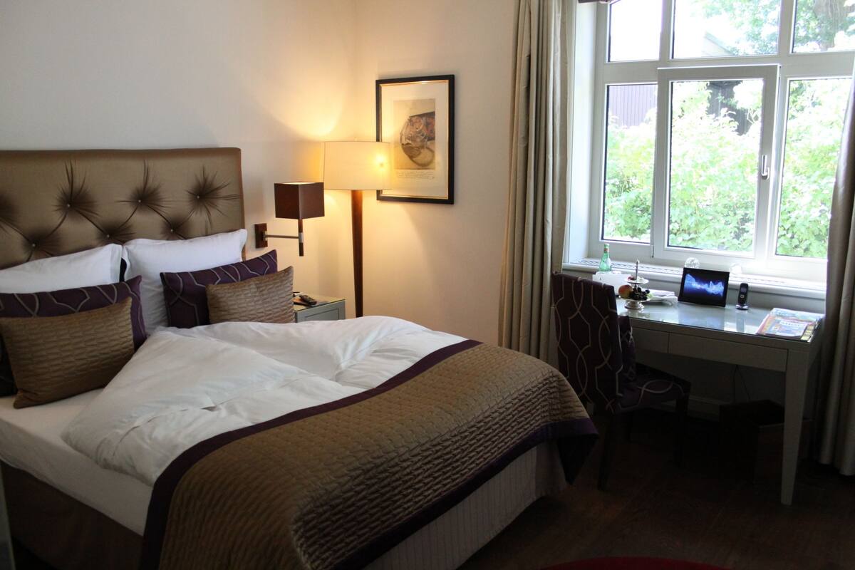 Quarto de hotel com uma cama grande, uma escrivaninha com um laptop, uma luminária e uma janela com vista para o verde.