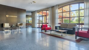 A hotel lobby with a modern design, featuring a reception desk, seating area with sofas and armchairs, large windows with views of the building and greenery, and a polished floor. - Hotel ILUNION Golf Badajoz (Badajoz)
