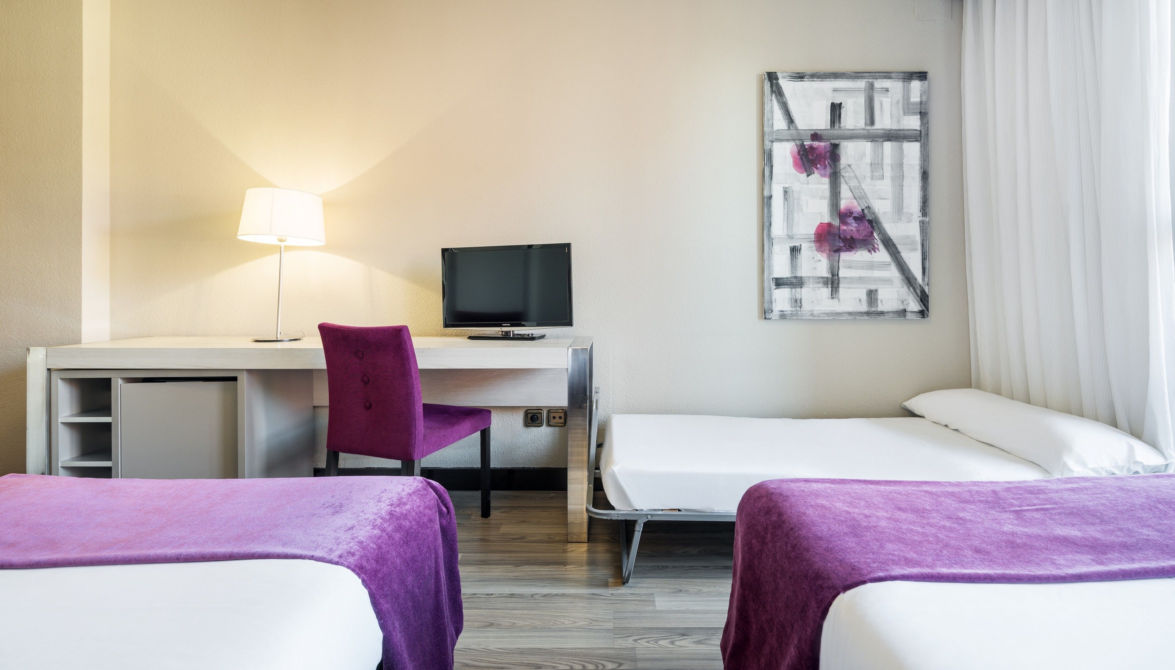 Habitación de hotel con escritorio, una silla, televisor, dos camas y un cuadro en la pared.