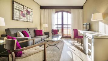 A modern hotel room with a grey sofa, a glass coffee table, a pink armchair, and a TV on a white desk.
