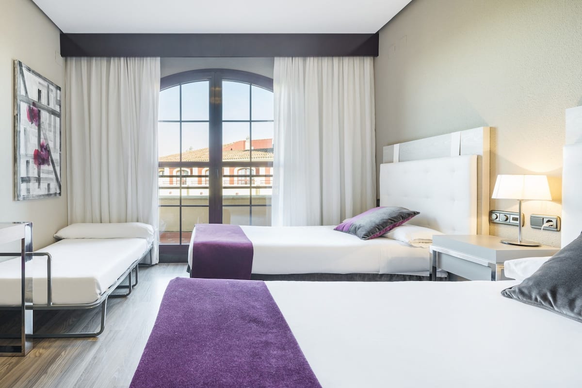 A hotel room with two beds, a desk, a chair, a TV, and a large window with curtains.