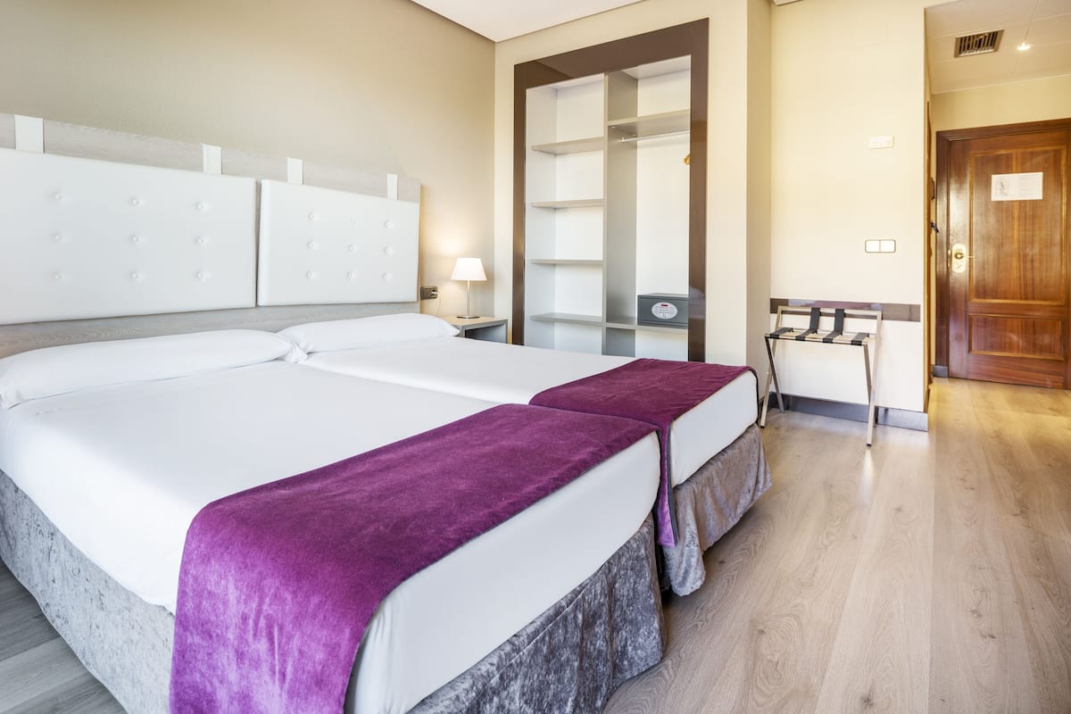 A double bed with a purple throw blanket, a nightstand, a bedside table, and a wooden door in a hotel room.