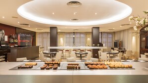 A modern hotel lobby with a central round table displaying pastries and a bar area with a selection of beverages. - Hotel ILUNION Golf Badajoz (Badajoz)