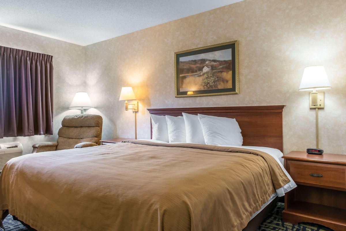 A hotel room with a large bed, two bedside lamps, a headboard, a painting on the wall, a chair, and a nightstand with a clock.