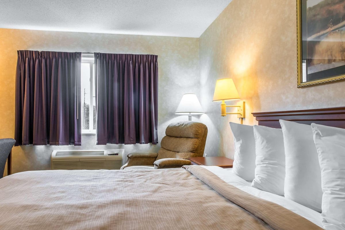 A hotel room with a bed, a chair, a lamp, and a window with curtains.