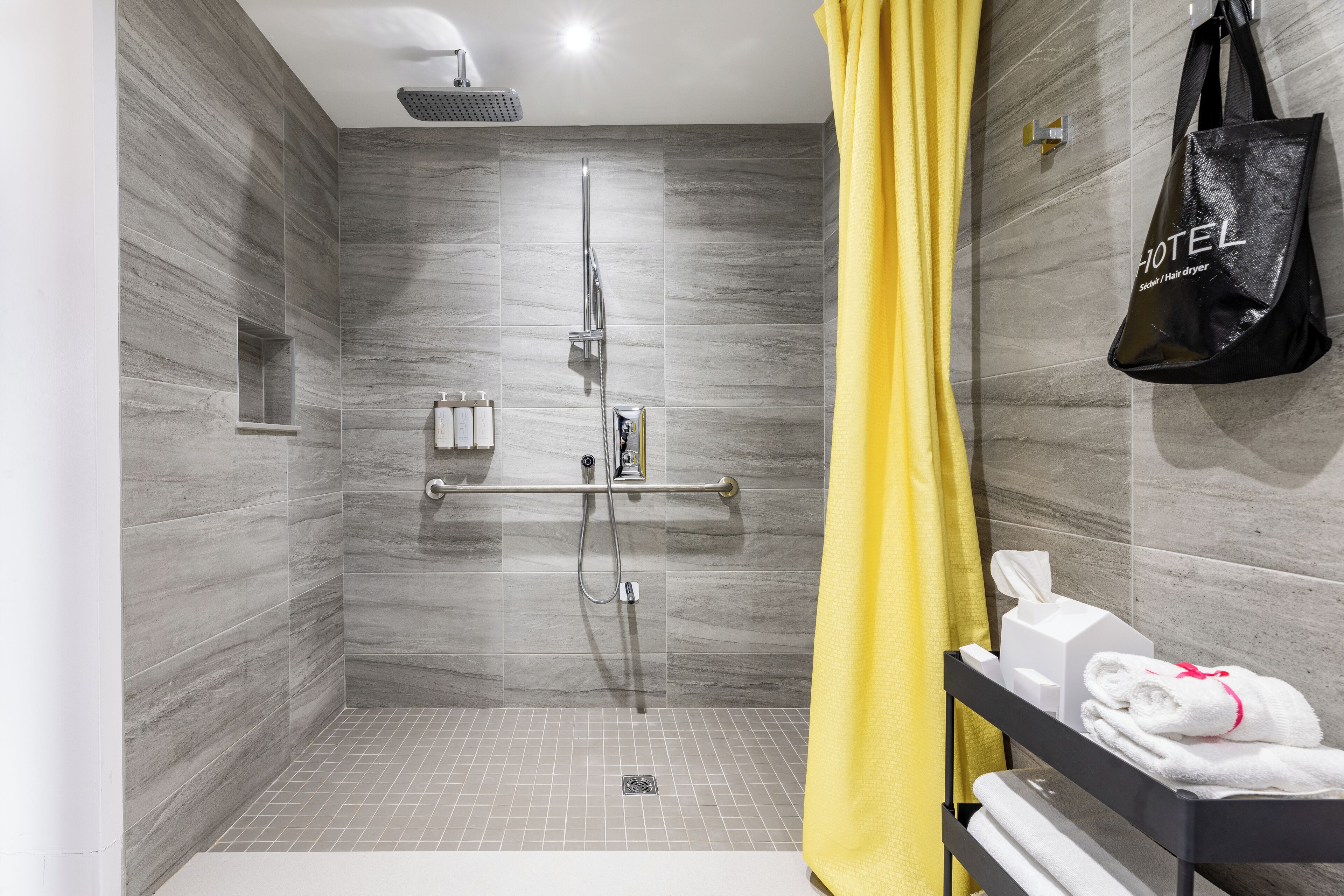 Een moderne douche ruimte met een glazen deur, een handdoekhouder met handdoeken en een zwarte tas met het logo van het hotel.