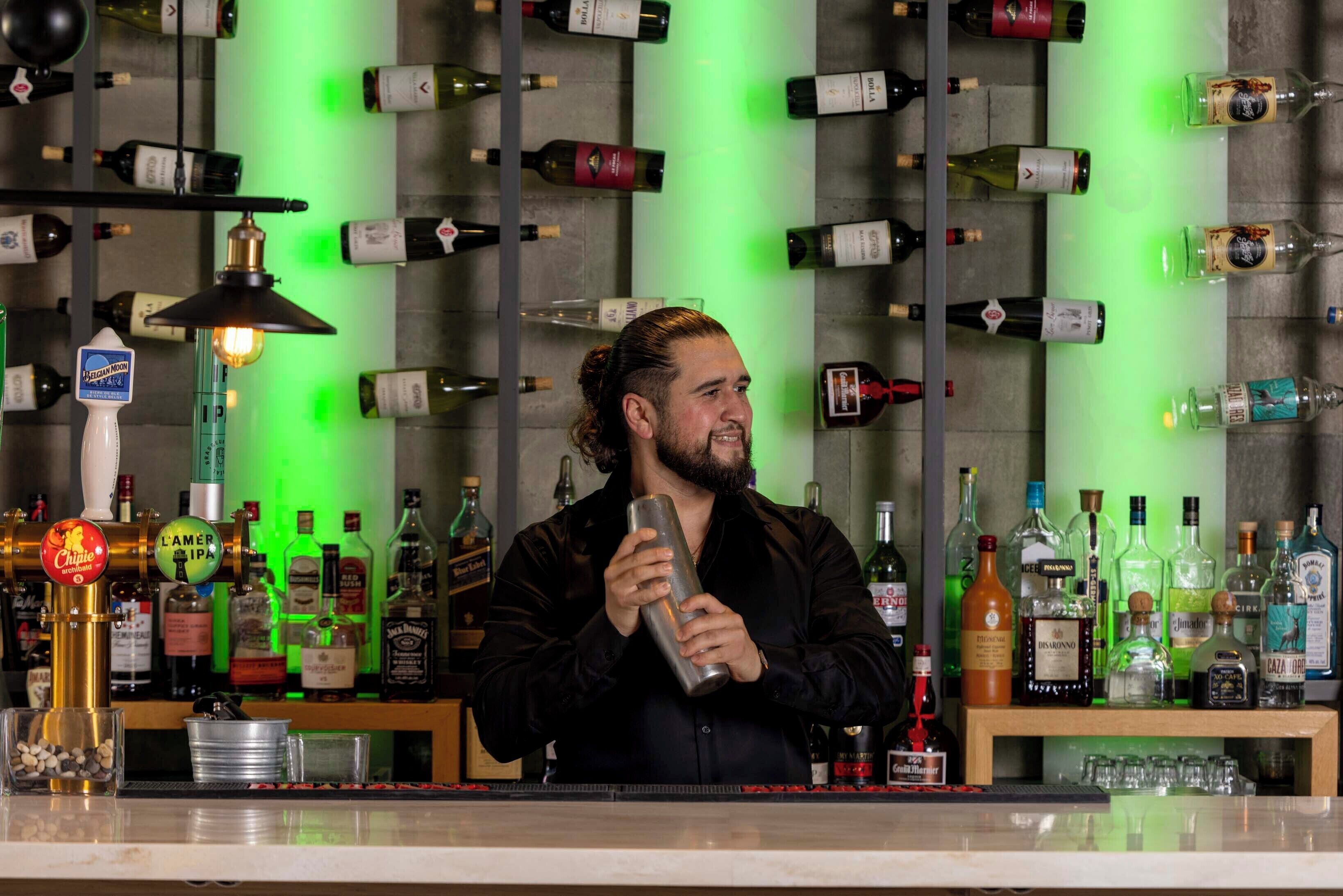 A bartender at a bar with a variety of liquor bottles and glasses on shelves behind him.