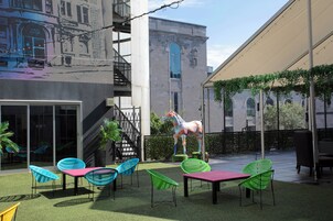 An outdoor patio with artificial grass, colorful chairs, a large giraffe sculpture, and a building with arched windows in the background.