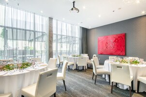 A dining area with round tables set for a meal, large windows, and a red abstract painting on the wall.