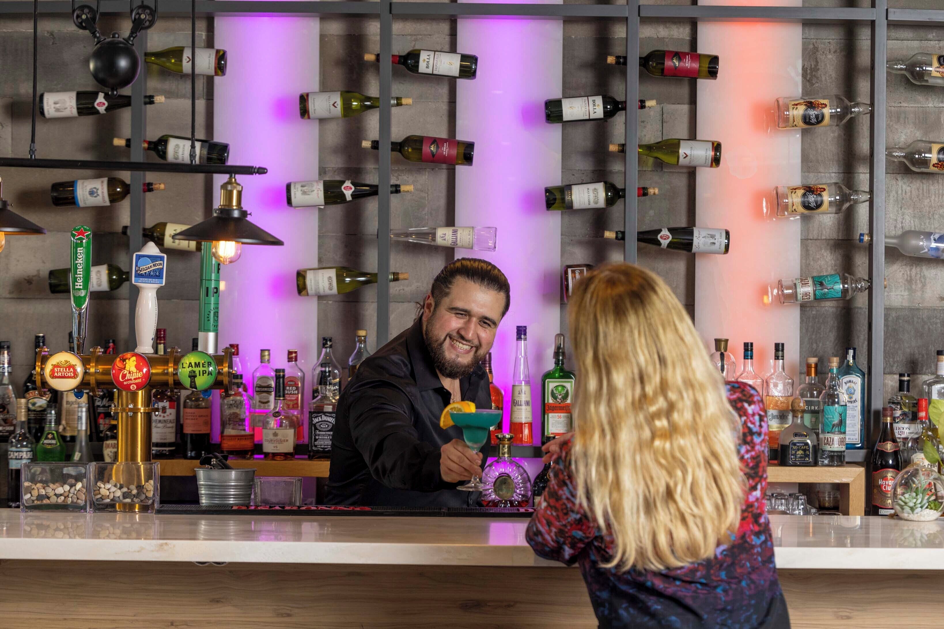 A bar area with a bartender and a customer. The bar has multiple liquor bottles and a variety of bar tools.