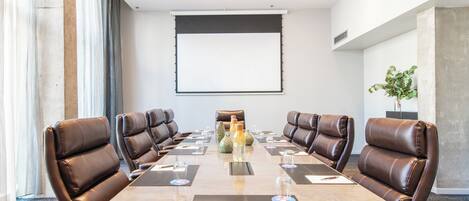 Un salón de conferencias con una larga mesa, sillas de cuero marrón, una pantalla de proyector y una planta en maceta.