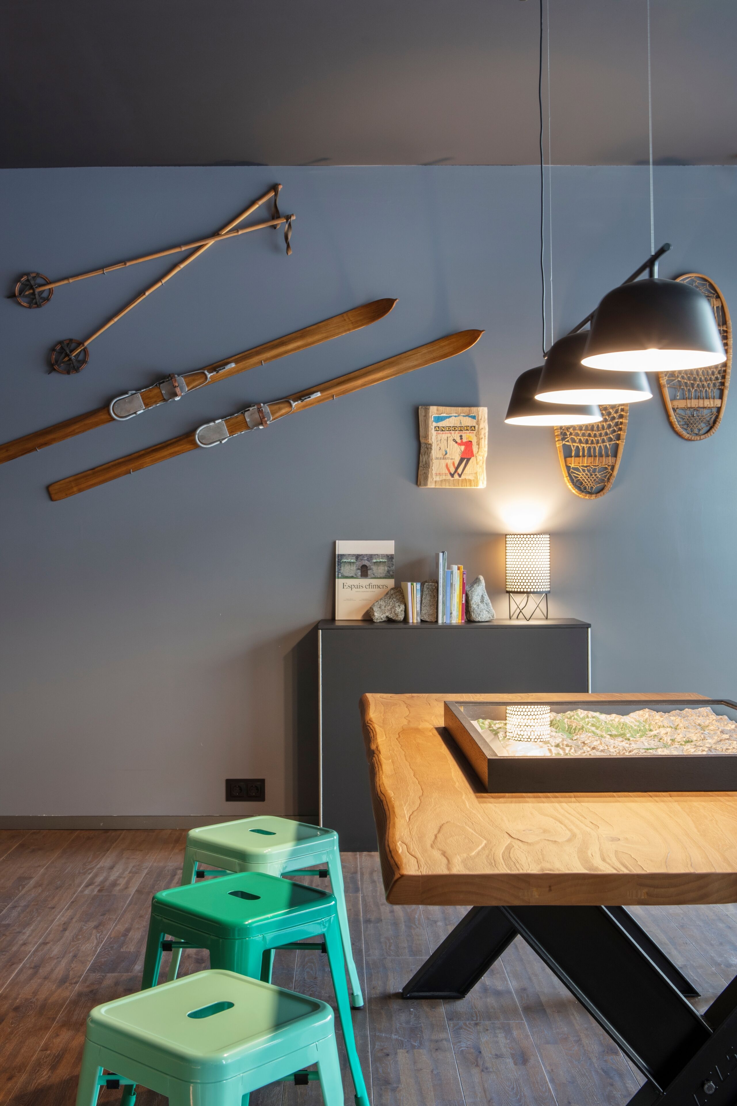 A room with a wooden table, green stools, skis on the wall, and a hanging light fixture.