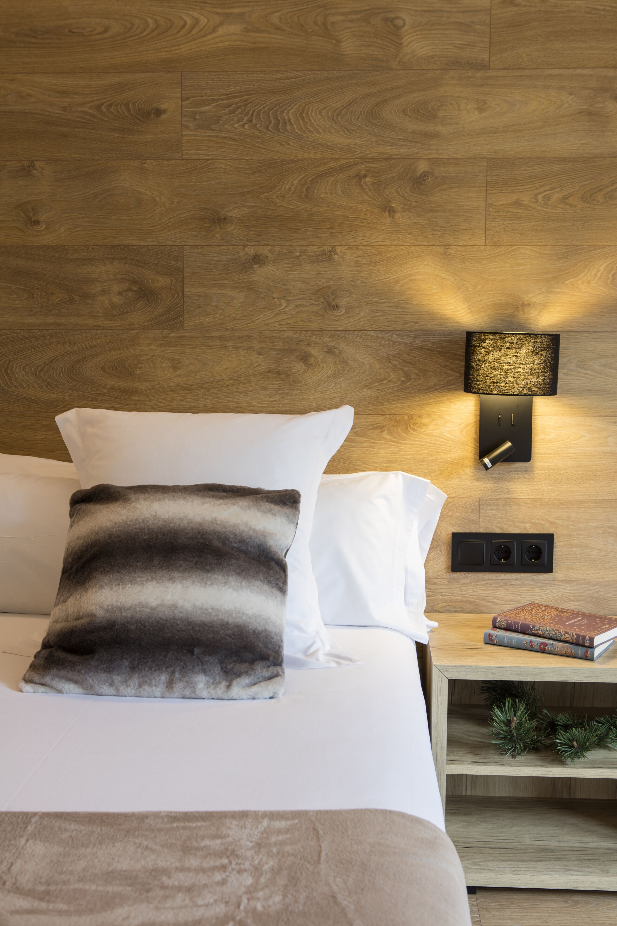 A neatly made bed with white and gray pillows, a wooden headboard, a wall-mounted light fixture, and a nightstand with books and a small plant.