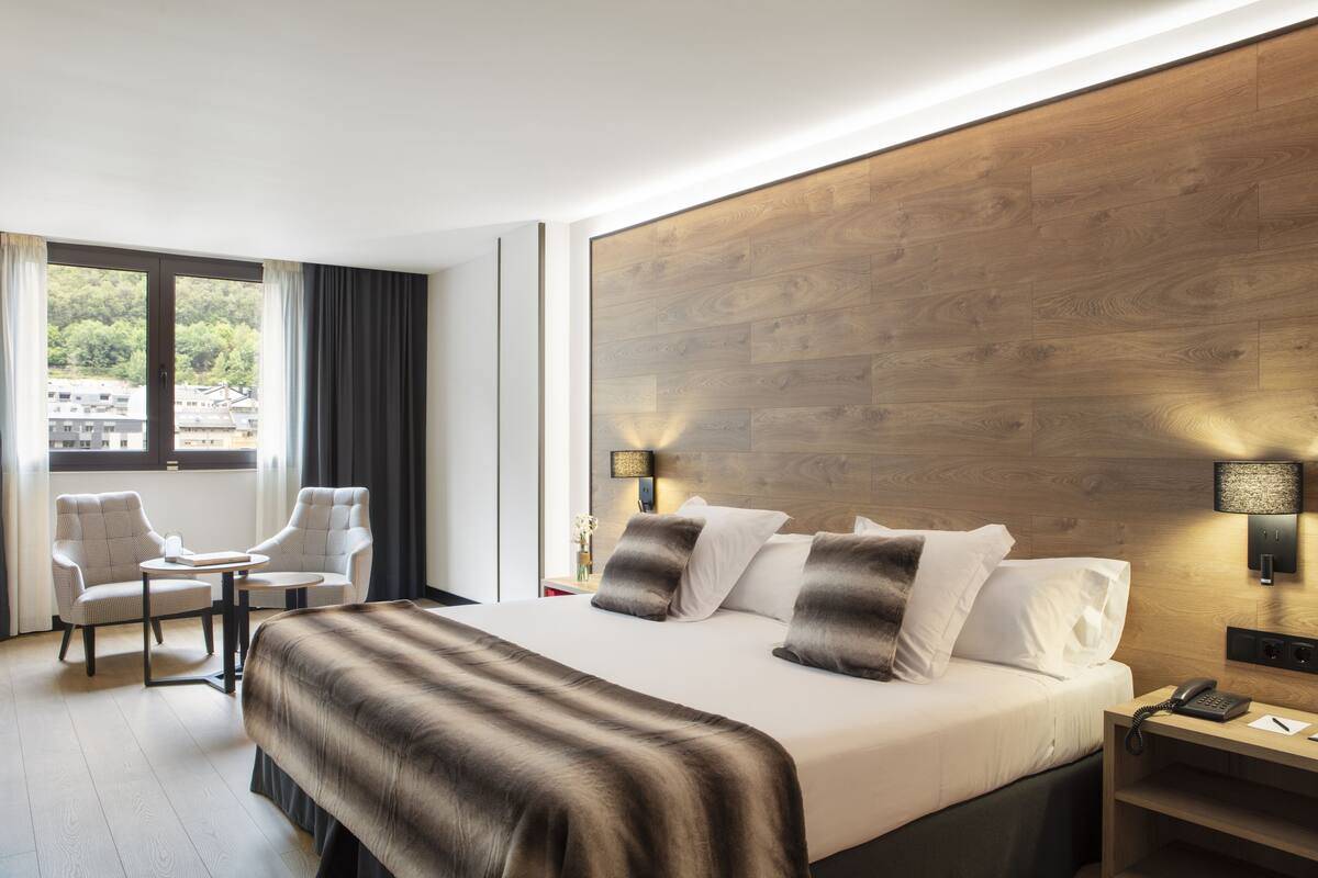 A modern hotel room with a large bed, two armchairs, a small table, a nightstand with a lamp, and a view of the city through large windows.