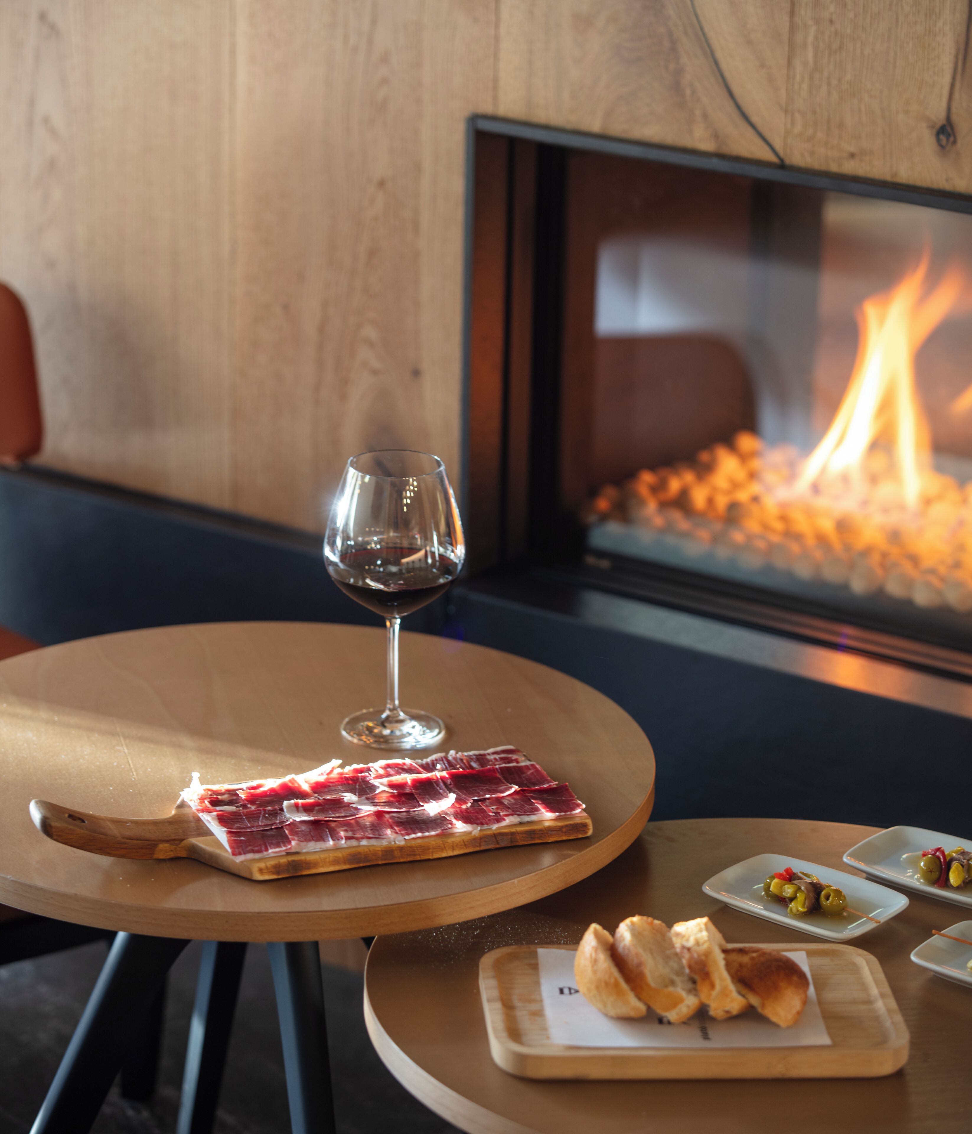 Una mesa de comedor con una copa de vino tinto, un plato de embutidos y un recipiente pequeño de aceitunas.