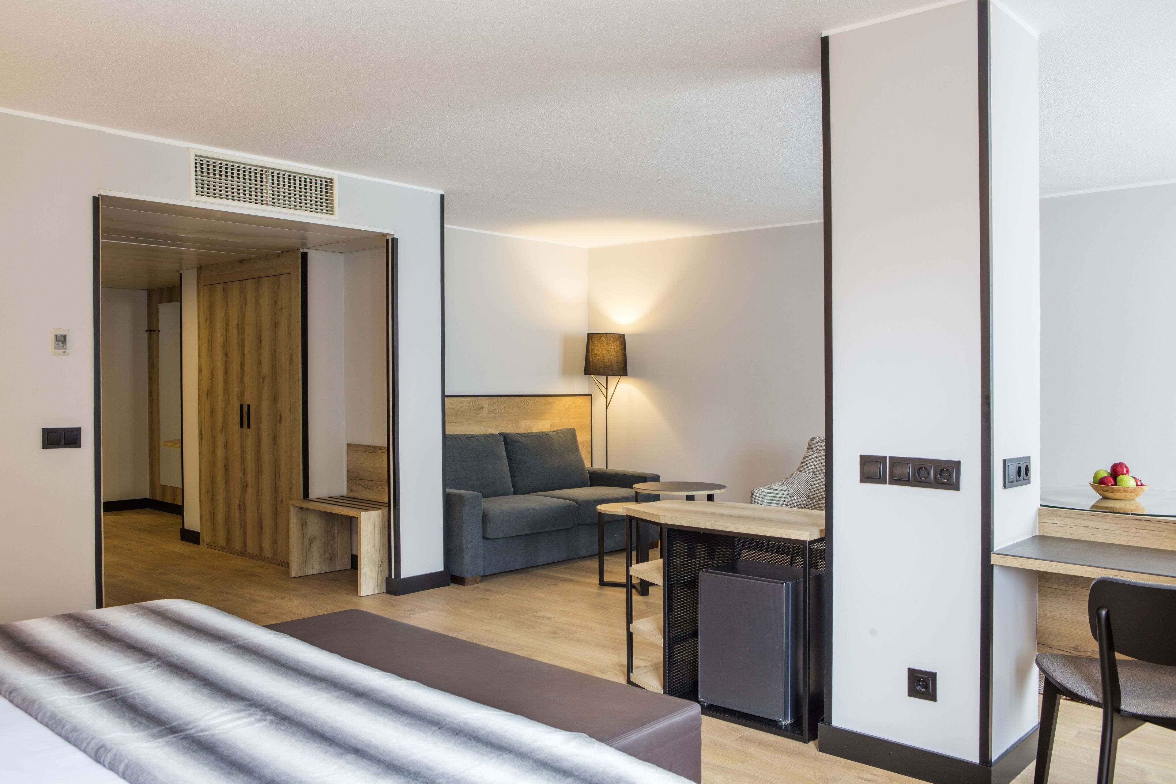 A modern hotel room with a bed, a sofa, a desk, and a small dining table. There is a lamp on the desk and a bowl of fruit on the table.